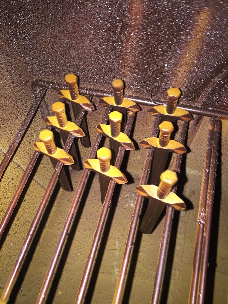 Nine miniture swords dangling between a metal rack being painted from yellow filler primer to a metallic bronze.